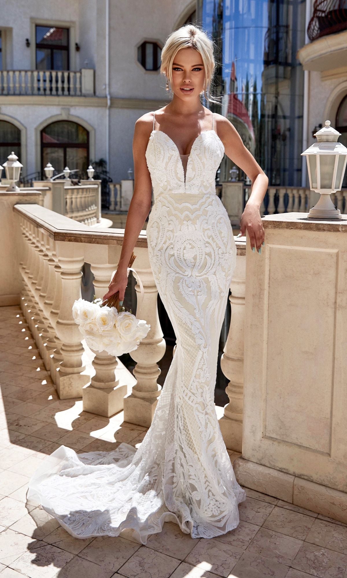 A woman in a fitted Long White Sequin Wedding Gown: Tina Holly T19280, featuring a mermaid silhouette and sweeping train, stands on a sunlit terrace holding a bouquet of white flowers.